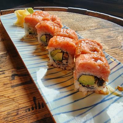 Sushi roll topped with seared pink fish and sauce, with a lemon slice