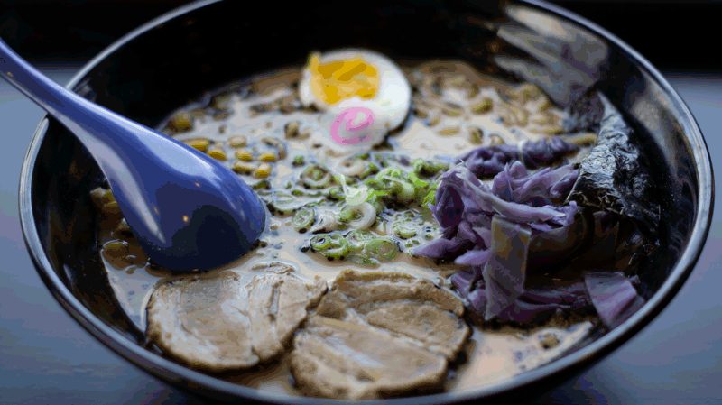 Tonkotsu ramen in a dark bowl with sliced pork, soft egg, scallions and a blue spoon